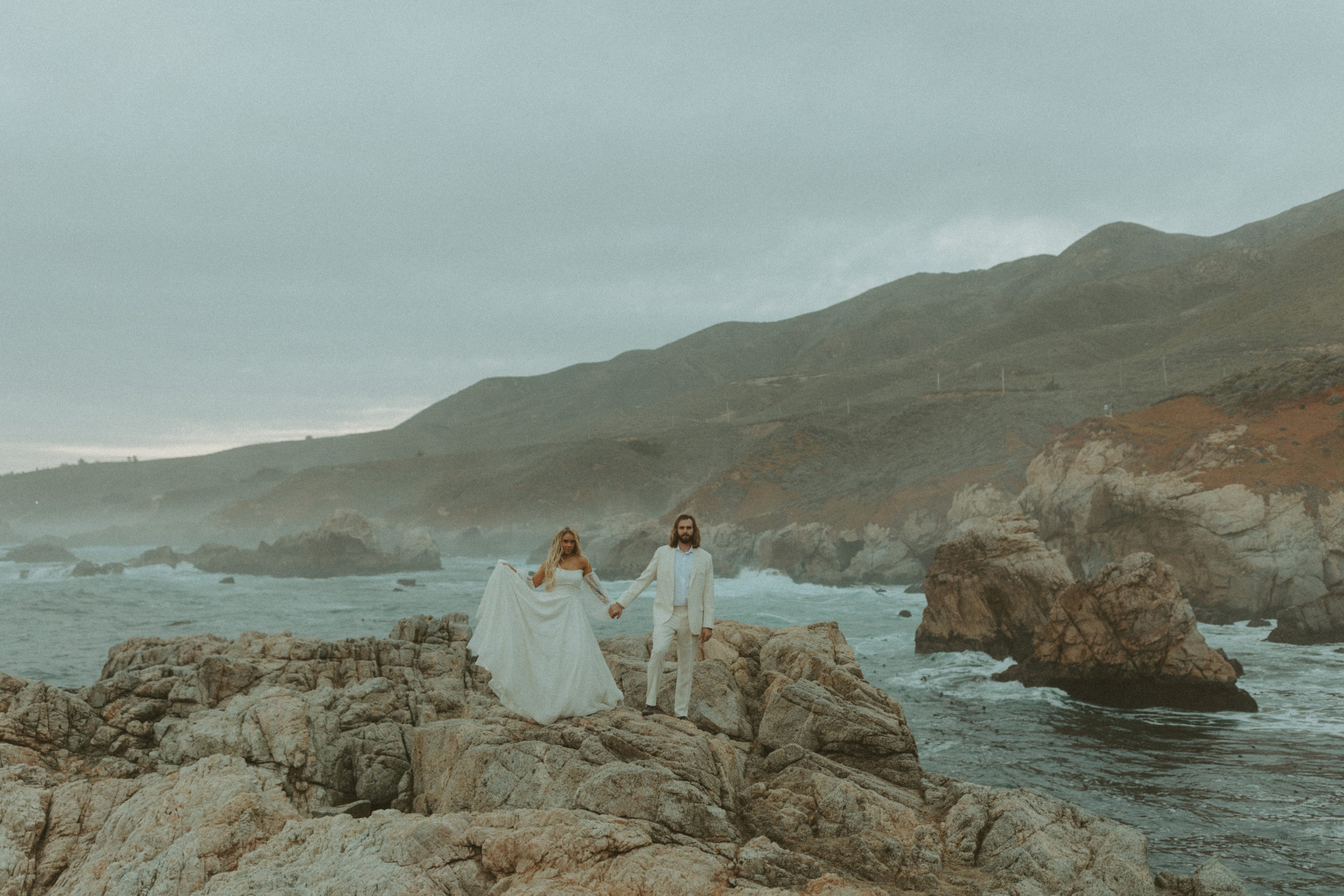 the couple holding hands and standing on the cliffs of Big Sur