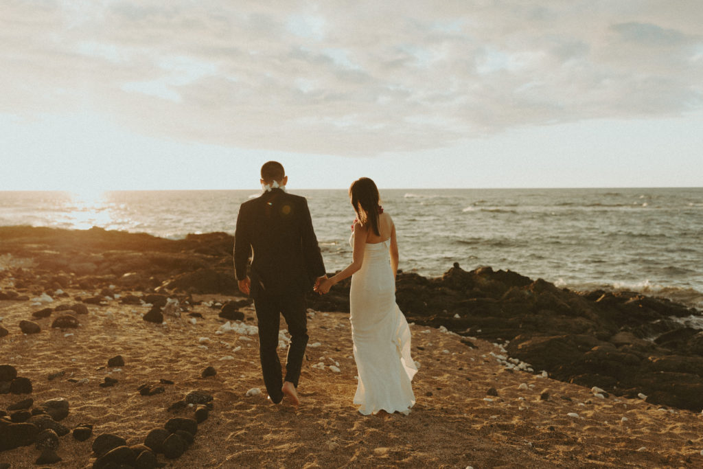 the wedding couple holding hands and walking along the beach in Hawaii