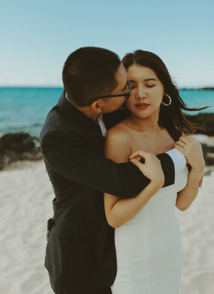 the groom kissing the bride's cheek on the beach in Hawaii