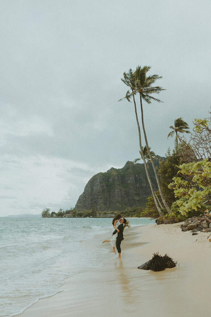 Bride and groom walking barefoot along Kaaawa Beach during golden hour
