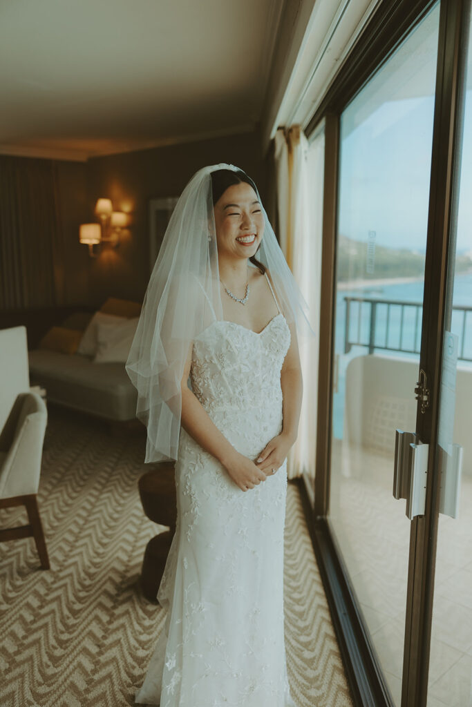 Bride smiling in a lace wedding dress and veil by the window. | Moana Surfrider
