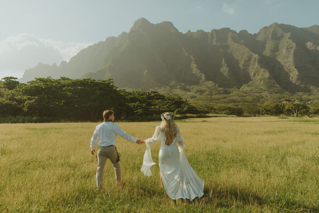 Bride and groom walking through Kualoa Regional Park with mountain views in the background.
