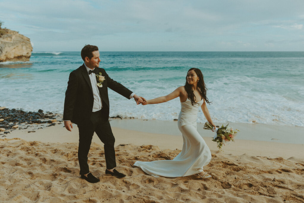 Bride and groom holding hands and smiling after their Shipwreck Beach elopement with ocean waves behind them
