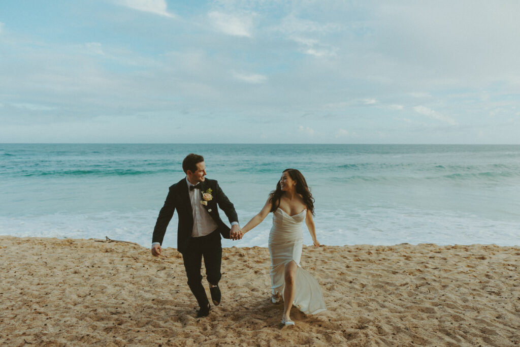 Bride and groom holding hands and smiling after their Shipwreck Beach elopement with ocean waves behind them
