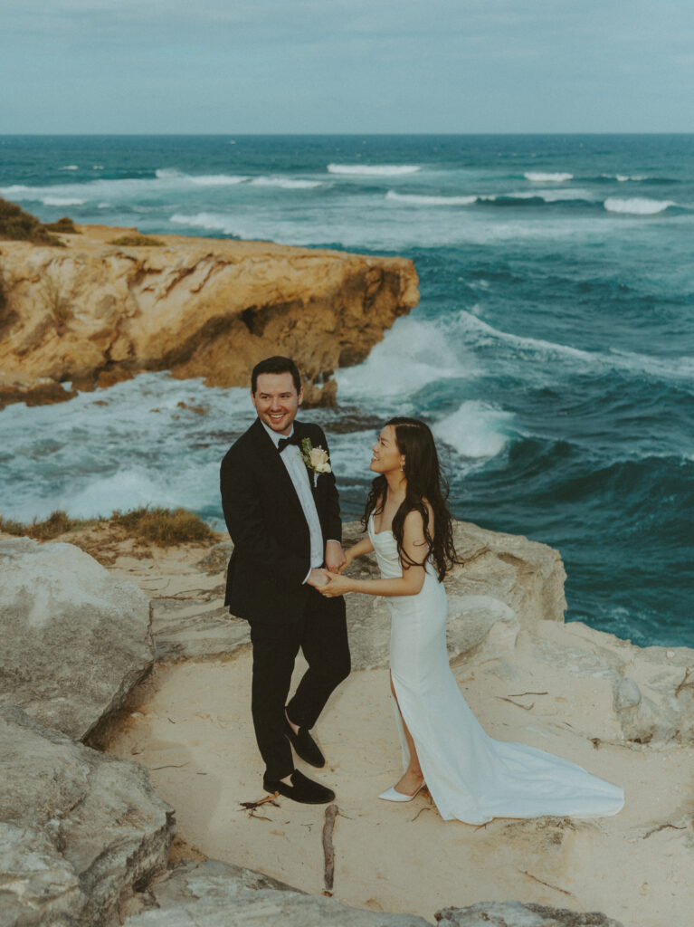 Bride and groom holding hands and smiling after their Shipwreck Beach elopement with ocean waves behind them
