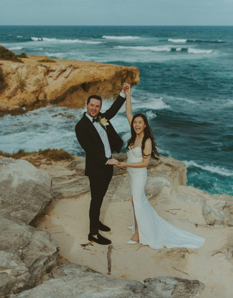 Bride and groom holding hands and smiling after their Shipwreck Beach elopement with ocean waves behind them
